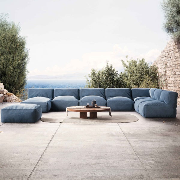 Modernes Outdoor-Lounge-Set mit blauem Modulsofa, Hocker und rundem Holz-Couchtisch auf einer Betonterrasse mit Meerblick.