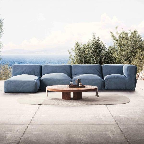Blaues modulares Lounge-Sofa auf einer hellen Steinterrasse mit Meerblick. Ein runder Holztisch steht auf einem ovalen Teppich.