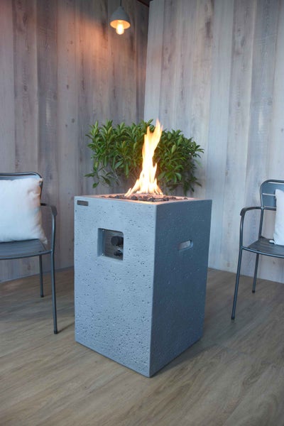 Quadratischer Gaskamin in Betonoptik mit lodernder Flamme auf einer Terrasse mit Metallstühlen und Holzwand.