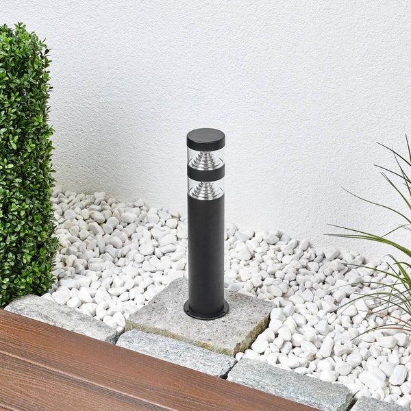 Moderne schwarze Pollerleuchte im Gartenbeet mit weißen Kieselsteinen neben einer grünen Hecke.