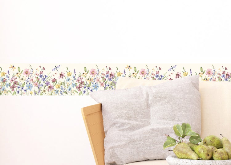 Selbstklebende Wandbordüre mit buntem Wildblumen- und Libellenmotiv an einer weißen Wand hinter einem Sessel mit Dekokissen.