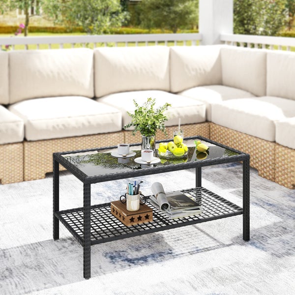 Rechteckiger Garten-Couchtisch aus schwarzem Polyrattan mit Glasplatte und zusätzlicher Ablage auf einer Terrasse vor einem hellen Ecksofa.