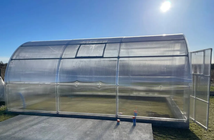 Großes Gewächshaus mit Hohlkammerplatten aus Polycarbonat, abgerundetem Dach, Metallrahmen, einem Dachfenster und einer offenen Tür.