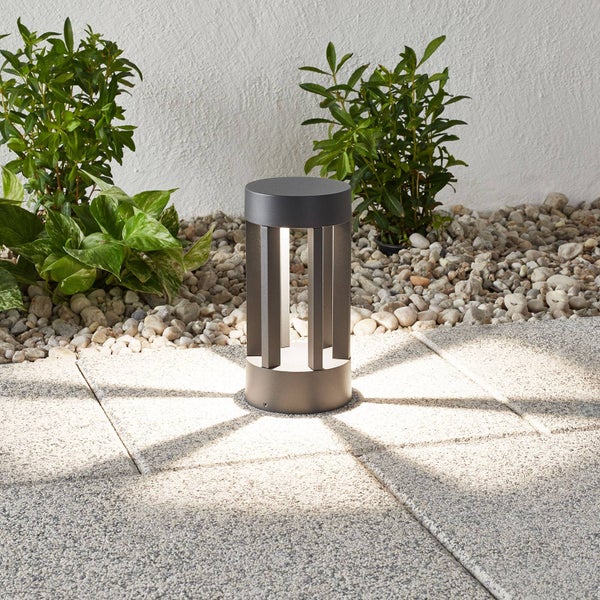 Moderne Pollerleuchte auf Steinplatten im Außenbereich, umgeben von Kiesbeeten und Pflanzen.