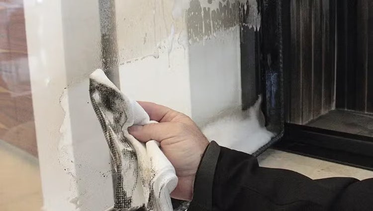 Eine Hand reinigt mit einem Tuch und Reinigungsschaum die verrußte Glasscheibe eines Kamins.