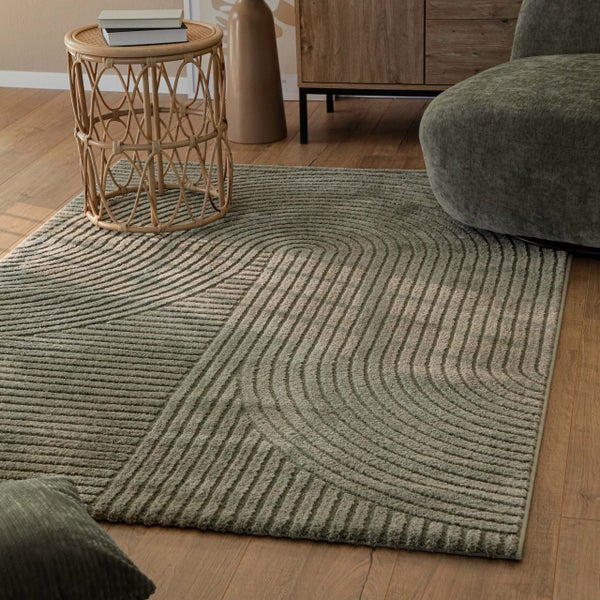 Rechteckiger Teppich mit geometrischer Hoch-Tief-Struktur in Salbeigrün auf Holzboden neben Rattanbeistelltisch und Sessel.