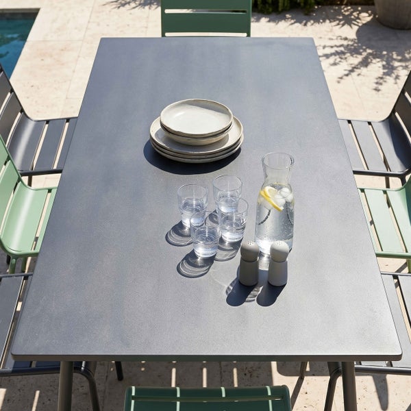 Rechteckiger Gartentisch mit Tellern, Gläsern und Karaffe, umgeben von Metallstühlen auf einer hellen Terrasse am Pool.