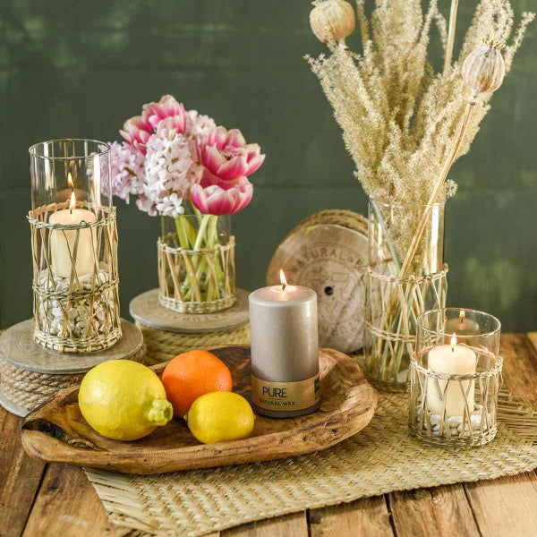 Dekorative Anordnung mit Glasvasen und Kerzenhaltern mit Geflecht, Stumpenkerzen, frischen Blumen, Trockengräsern und Zitrusfrüchten auf einem Holztisch.