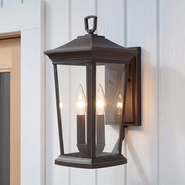 Klassische Außenwandleuchte in Laternenform aus Metall mit zwei brennenden Kerzenlampen, montiert an einer Holzwand.