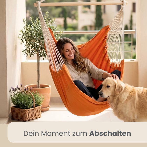 Eine Frau entspannt in einem Hängesessel auf einem hellen Balkon mit einem Hund, einem Olivenbaum im Topf und einem Kräuterkorb.