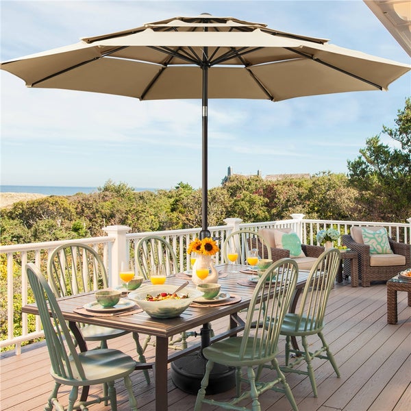 Beiger, dreistufiger Marktschirm auf einer Holzterrasse mit Esstisch und grünen Stühlen, mit Blick auf die Küste.
