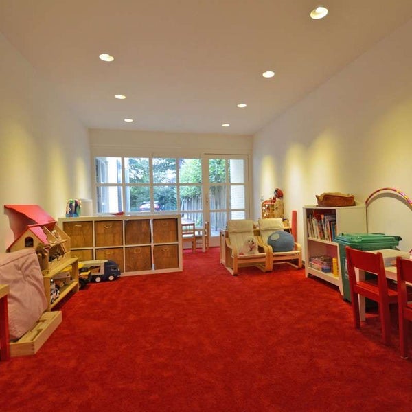 Helles Spielzimmer mit rotem Teppichboden, weißen Wänden, runden Deckeneinbaustrahlern, Spielzeugregalen, Kindermöbeln und großer Fensterfront.