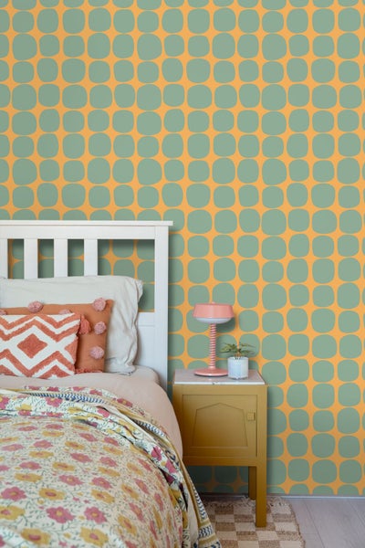 Vliestapete mit Retromuster in einem hellen Schlafzimmer mit weißem Holzbett, floraler Bettwäsche und gelbem Nachttisch.