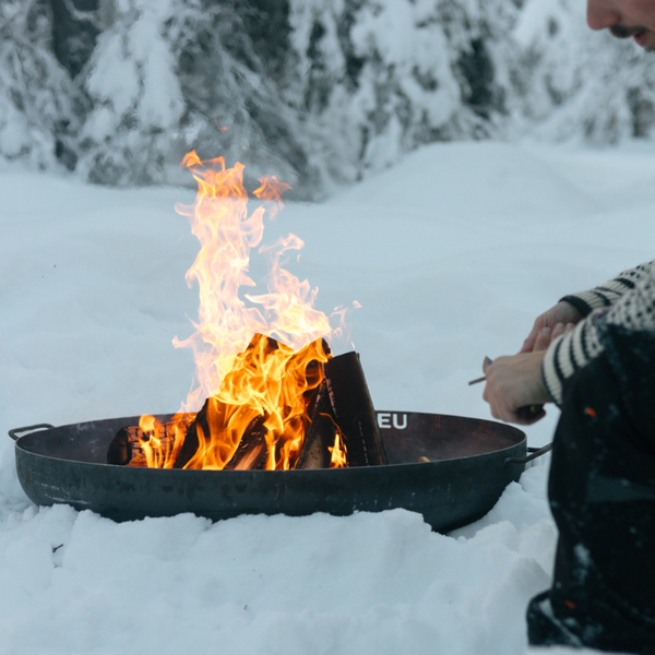 Feuerschale aus Metall mit brennendem Holz in einer verschneiten Winterlandschaft.