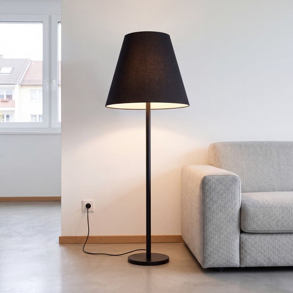 Moderne Stehleuchte mit schwarzem Textilschirm und schwarzem Metallgestell neben einem hellgrauen Sofa in einem hellen Wohnzimmer.