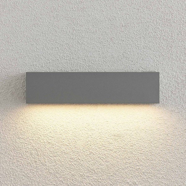 Rechteckige Außenwandleuchte in Dunkelgrau mit warmweißem Lichtschein nach unten auf einer strukturierten Wand.