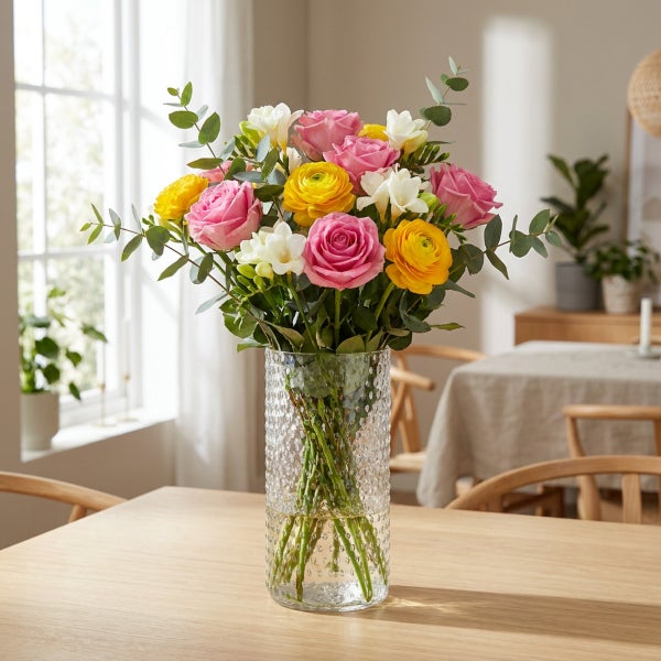 Bunter Blumenstrauß aus Rosen, Ranunkeln und Fresien in einer strukturierten Glasvase auf einem Holztisch in einem hellen Wohnzimmer.