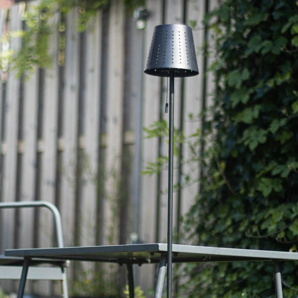 Moderne Außenstehleuchte mit perforiertem Schirm und Zugschalter auf einer Terrasse neben einem Gartentisch.
