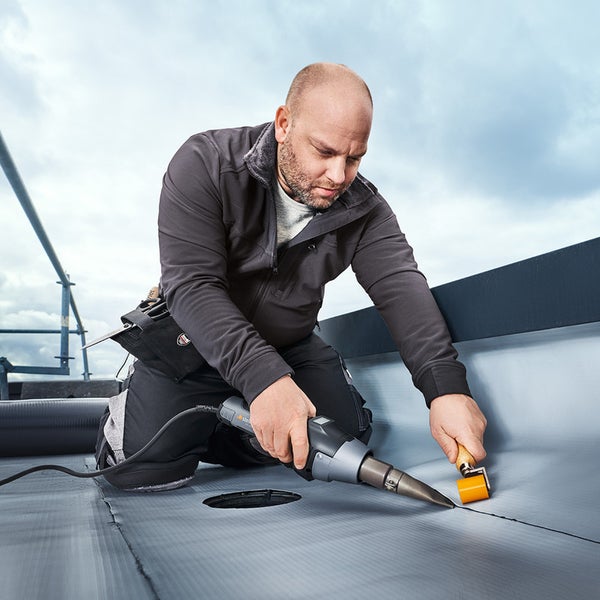 Handwerker verschweißt Dachbahnen mit einem Heißluftgebläse und einer Andrückrolle auf einem Flachdach.