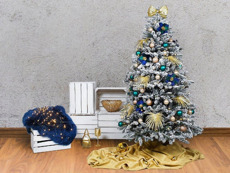 Geschmückter, künstlicher Weihnachtsbaum mit Kunstschnee, Kugeln und Dekoration neben weißen Holzkisten und einem beleuchteten Fell auf Holzboden.