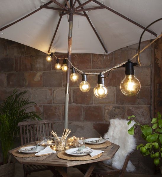 Gedeckter Tisch mit Sonnenschirm und Lichterkette im Freien.