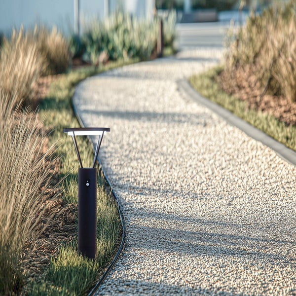 Moderne Sockelleuchte mit Sensor an einem hellen, geschwungenen Kiesweg im Garten mit Gräsern.