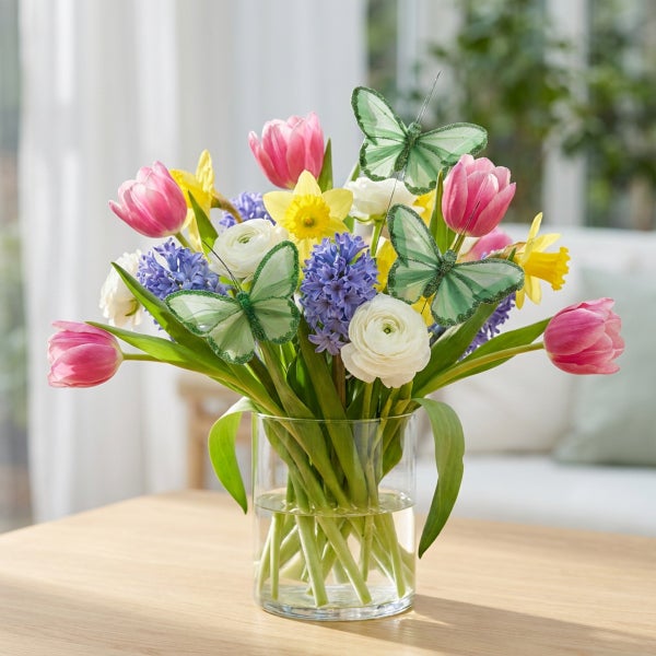 Frühlingsblumenstrauß mit Tulpen, Narzissen, Hyazinthen und Ranunkeln in einer Glasvase, dekoriert mit zwei künstlichen Schmetterlingen.