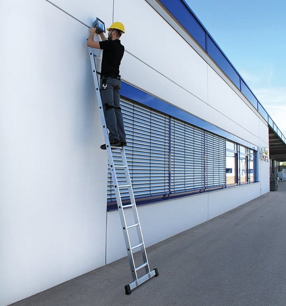 Handwerker mit Helm auf einer Aluminium Anlegeleiter bei der Montage eines Wandstrahlers an einer Fassade.