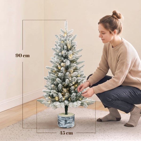 Eine Frau dekoriert einen 90 Zentimeter hohen, künstlichen, beschneiten Weihnachtsbaum mit integrierter LED Beleuchtung in einem dekorativen Topf.