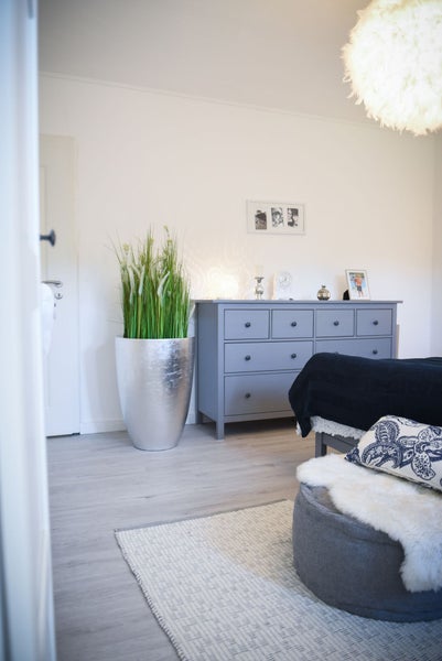 Modernes Schlafzimmer mit grauer Kommode, silbernem Pflanzgefäß mit Kunstgras und einem grauen Hocker mit weißem Fell.