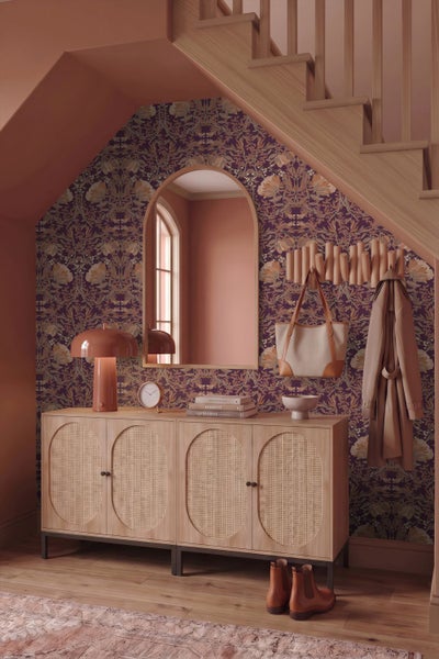 Hell gestalteter Flur mit einem Sideboard aus Holz mit Rattantüren, einem Rundbogenspiegel und floraler Tapete. Inklusive Treppe und Garderobe.