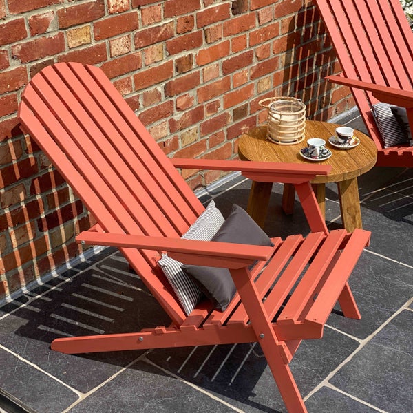 Zwei terrakottafarbene Adirondack-Gartenstühle aus Holz mit Kissen auf einer Terrasse neben einem hölzernen Beistelltisch vor einer Ziegelwand.
