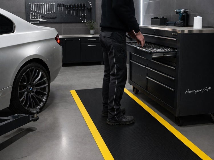 Eine Person steht auf einer Anti-Ermüdungsmatte mit gelben Rändern vor einem Werkstattwagen in einer organisierten Garage.