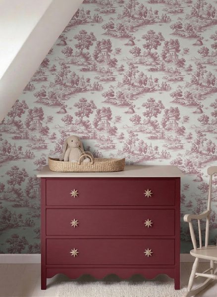 Heller Raum mit Toile-de-Jouy-Mustertapete, einer weinroten Kommode mit drei Schubladen und Sternknöpfen sowie einem hölzernen Schaukelstuhl.