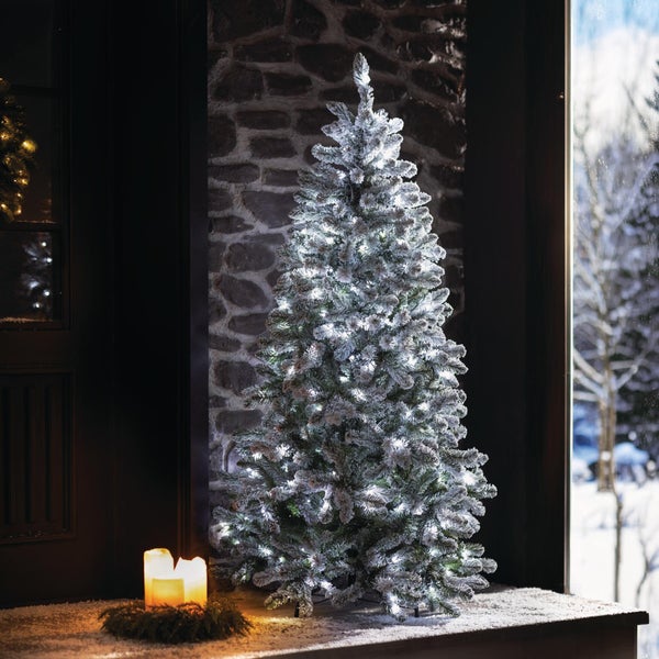 Künstlicher, verschneiter Weihnachtsbaum mit integrierter LED Beleuchtung vor einer Steinwand.