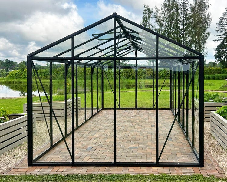 Großzügiges modernes Gewächshaus mit schwarzem Metallrahmen und Klarglas auf gepflastertem Boden in einem hellen Garten.