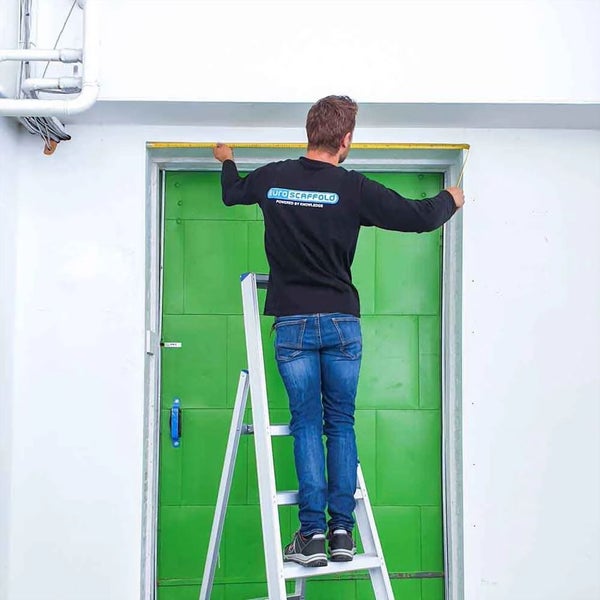 Mann auf Leiter misst mit einem Maßband eine Türöffnung.