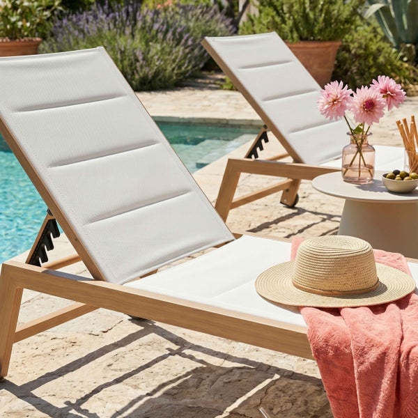 Zwei moderne Gartenliegen aus Holz mit weißer Textilbespannung auf einer Steinterrasse am Pool mit Hut und Handtuch.