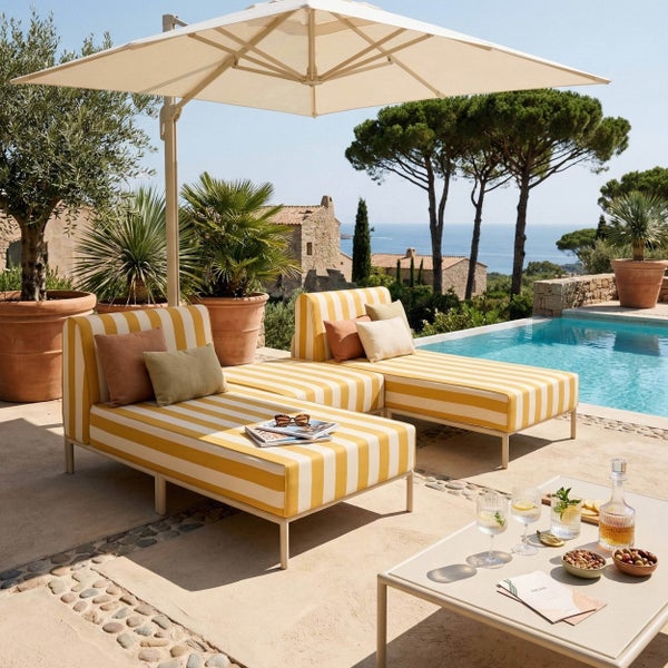 Helle mediterrane Terrasse mit zwei gestreiften Sonnenliegen unter einem großen weißen Sonnenschirm neben einem Pool mit Meerblick.