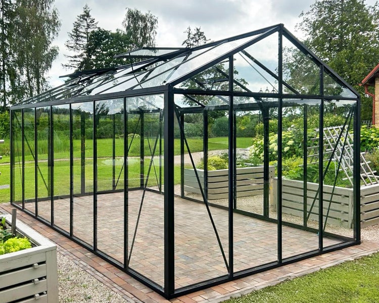 Großes Gewächshaus aus Glas mit schwarzem Metallrahmen und geöffneten Dachfenstern auf gepflasterter Fläche in einem Garten.
