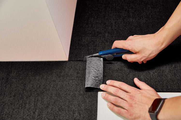 Eine Person schneidet einen dunklen, strukturierten Bodenbelag mit einem blauen Cuttermesser an einer Wand entlang zu.