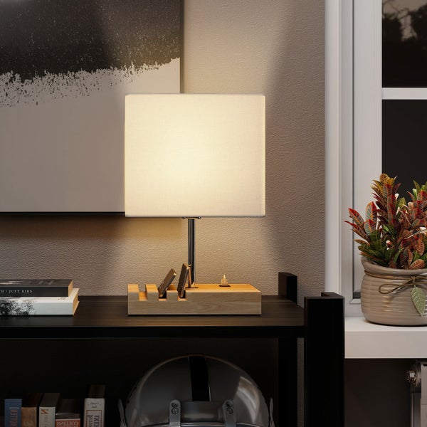 Moderne Tischleuchte mit quadratischem Textilschirm und Holzsockel inklusive Mobiltelefon-Halterung und Ladeanschluss auf einem Regal.