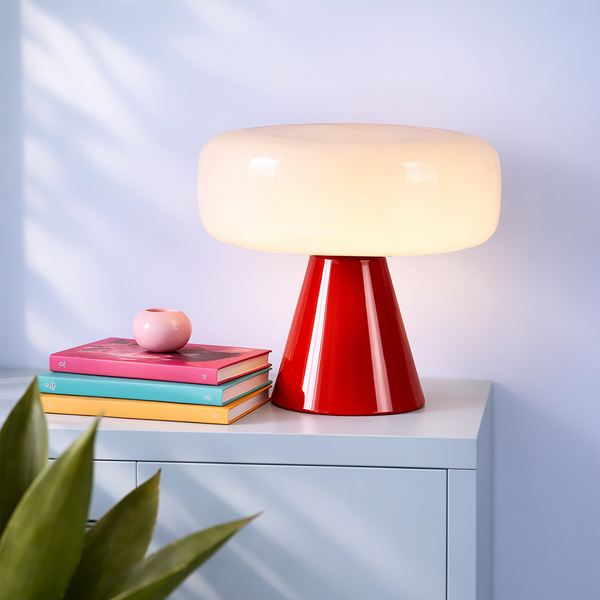 Moderne Tischleuchte mit weißem Glasschirm und rotem konischem Fuß auf einer hellen Kommode neben bunten Büchern in einem hellen Raum.