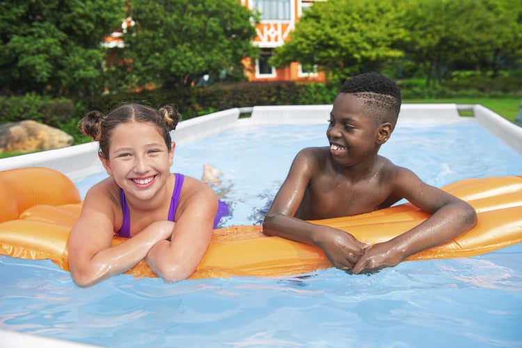 Zwei Kinder spielen in einem hellen, oberirdischen Rahmenpool auf einer orangefarbenen Luftmatratze in einem Garten.