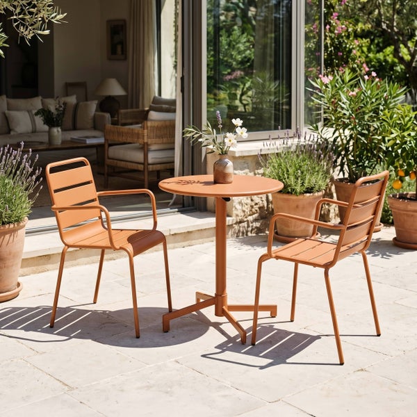 Moderne terrakottafarbene Gartenmöbel aus Metall mit rundem Tisch und zwei Stühlen auf einer hellen Steinterrasse mit Pflanzen.