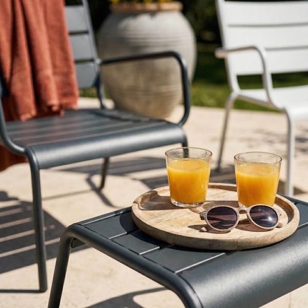 Moderne Gartenmöbel aus Metall mit Stühlen und Beistelltisch. Holztablett mit Gläsern und Sonnenbrille auf dem Tisch im hellen Außenbereich.