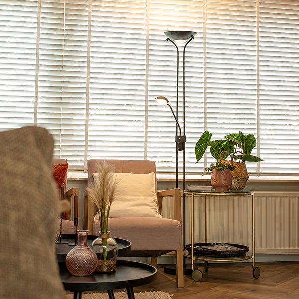 Wohnzimmer mit Sessel, Tisch und Stehlampe vor einem Fenster mit Jalousien