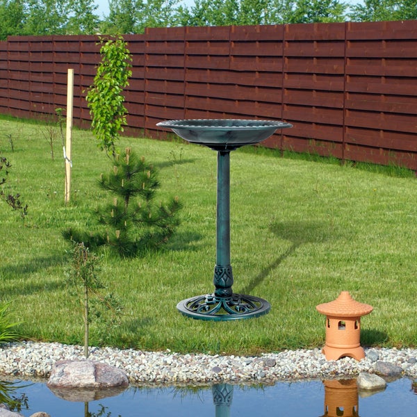 Klassische Vogeltränke auf einem verzierten Standfuß in einem gepflegten Garten mit Rasen, Holzschutzzaun und kleinem Teich.