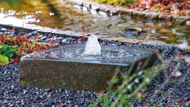 Quadratischer Gartenbrunnen aus Stein mit Wasserspiel in einem Kiesbeet vor einem Teich.