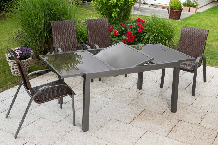 Ausziehbarer Gartentisch mit Glasplatte und vier Gartenstühlen aus Polyrattan auf einer gepflasterten Terrasse im Garten.